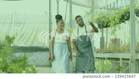 Walking man in apron and customer wearing light blue skirt inside greenhouse, with hanging plants Walking man in apron and customer wearing light blue skirt inside greenhouse, with hanging plants 139425647