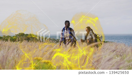 Hiking couple wearing jackets walking through tall dune grass by ocean, with packs poles sleep pad Hiking couple wearing jackets walking through tall dune grass by ocean, with packs poles sleep pad 139425650