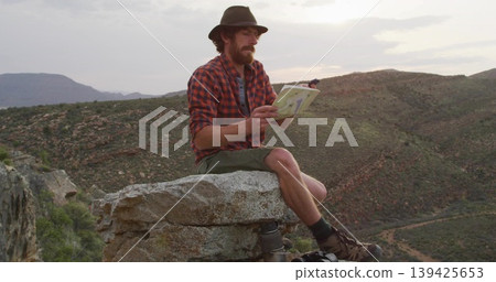 Examining paper map, bearded hiker wearing fedora sitting on canyon rim, with backpack, metal stove Examining paper map, bearded hiker wearing fedora sitting on canyon rim, with backpack, metal stove 139425653