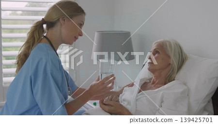 Leaning nurse wearing blue scrubs offering glass of water to patient on hospital bed in room 139425701