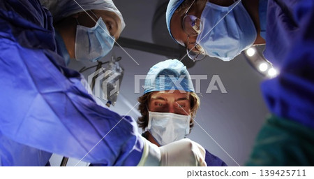 Male surgeon in blue scrubs leaning over operating table in operating room, with scalpel, IV bag Male surgeon in blue scrubs leaning over operating table in operating room, with scalpel, IV bag 139425711