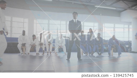 Standing man in black suit officiating martial arts session on interlocking mats in gym, uniforms Standing man in black suit officiating martial arts session on interlocking mats in gym, uniforms 139425745