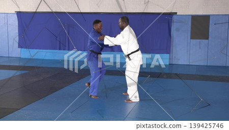 Two judokas practicing judo in dojo, with blue white gis black belts interlocking mats padded walls 139425746