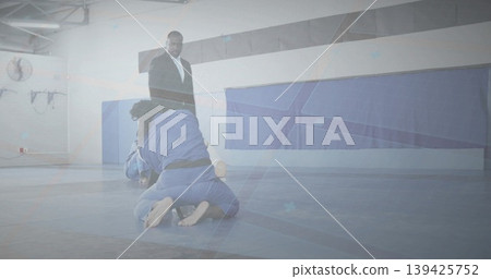 Grappling opponents in blue gis exchanging ground holds on martial arts gym mat, with ring ropes Grappling opponents in blue gis exchanging ground holds on martial arts gym mat, with ring ropes 139425752
