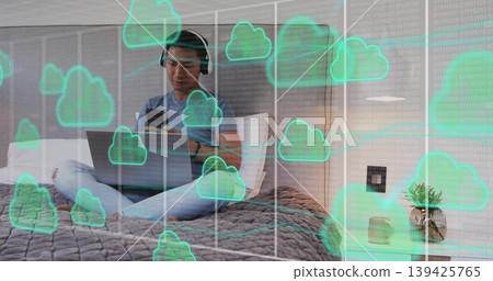 Sitting Asian man wearing headphones tapping smartphone and coding on laptop on bed, cloud overlay 139425765