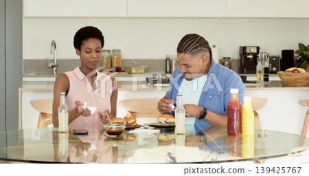Sharing burgers and dipping sauce with spoon, couple wiping with napkin in modern home kitchen Sharing burgers and dipping sauce with spoon, couple wiping with napkin in modern home kitchen 139425767
