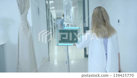 Walking patient with wristband in gown pushing IV drip stand down white corridor, sterile ward 139425770
