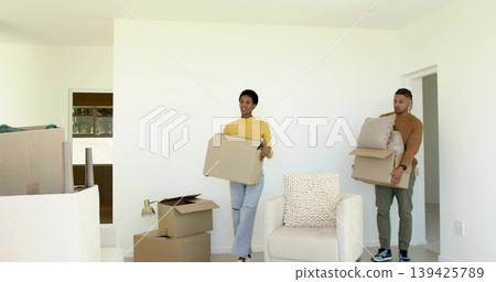 Couple wearing casual clothes carrying moving boxes in living room with wrapped furniture 139425789
