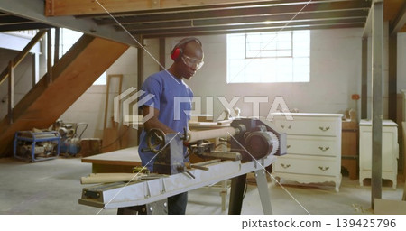 Mid adult male carpenter shaping spindle on lathe in woodshop, with goggles and earmuffs Mid adult male carpenter shaping spindle on lathe in woodshop, with goggles and earmuffs 139425796