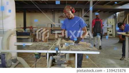 Operating carpenter feeding wooden board through table saw in workshop, with sawdust and gloves Operating carpenter feeding wooden board through table saw in workshop, with sawdust and gloves 139425876