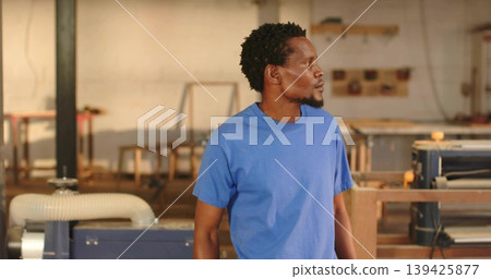 Standing woodworker wearing blue t-shirt and jeans in workshop with machines, benches, tool shelves Standing woodworker wearing blue t-shirt and jeans in workshop with machines, benches, tool shelves 139425877