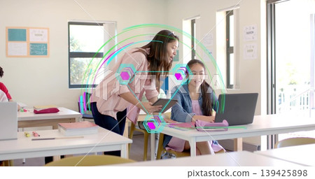 Explaining instructor leaning over student working on laptop in classroom, with tablet guidance Explaining instructor leaning over student working on laptop in classroom, with tablet guidance 139425898