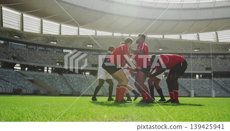 Crouching rugby players in red jerseys forming scrum in stadium with goal posts, referee whistle Crouching rugby players in red jerseys forming scrum in stadium with goal posts, referee whistle 139425941