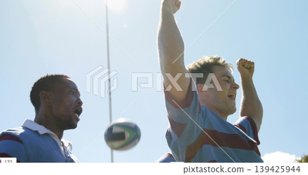 Cheering players wearing blue maroon striped jerseys on rugby pitch, with rugby ball and goal post 139425944