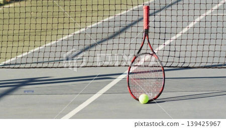 Black and red tennis racket leaning against net in lined hard court, with yellow tennis ball 139425967