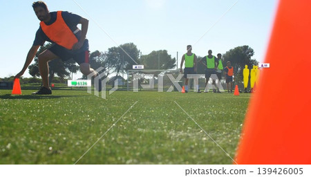 Lunging male athlete in orange bib reaching cone on turf, with goal, mannequins, fence, houses Lunging male athlete in orange bib reaching cone on turf, with goal, mannequins, fence, houses 139426005