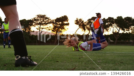 Lying blonde player grimacing on grass pitch at sunset, wearing neon bib, sports kit, cleats Lying blonde player grimacing on grass pitch at sunset, wearing neon bib, sports kit, cleats 139426016