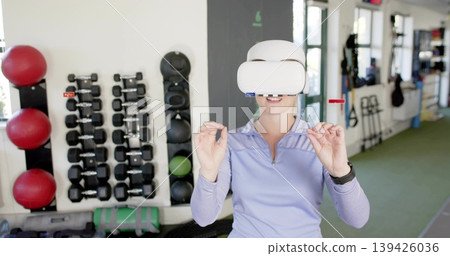 Standing woman in purple top and white VR headset interacting in studio, dumbbell rack, red balls Standing woman in purple top and white VR headset interacting in studio, dumbbell rack, red balls 139426036