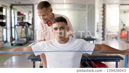 Holding blue resistance band, white shirt exerciser pulling across chest in gym, trainer guiding 139426043
