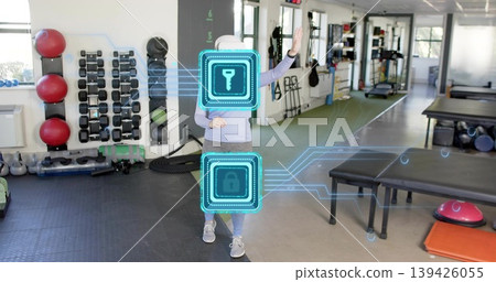 Standing adult woman gesturing toward AR holograms at gym, wearing white cap and gray leggings Standing adult woman gesturing toward AR holograms at gym, wearing white cap and gray leggings 139426055