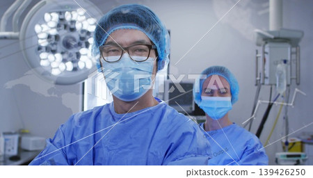 Standing surgeons wearing blue scrubs with caps in operating room, with surgical light and IV stand 139426250