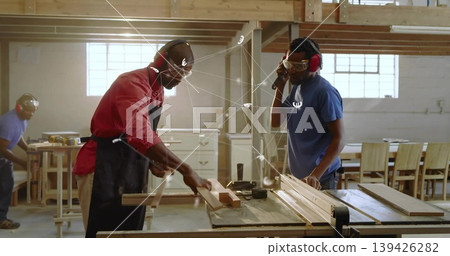Two carpenters with safety goggles and ear protection cutting wooden board on table saw in workshop Two carpenters with safety goggles and ear protection cutting wooden board on table saw in workshop 139426282