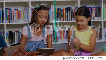 Sitting two girls in denim overalls, yellow bow top reading tablets in library, with bookshelves 139426295