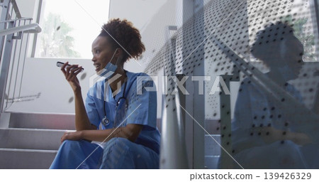 Sitting nurse in scrubs speaking into smartphone in stairwell, with surgical mask and stethoscope Sitting nurse in scrubs speaking into smartphone in stairwell, with surgical mask and stethoscope 139426329