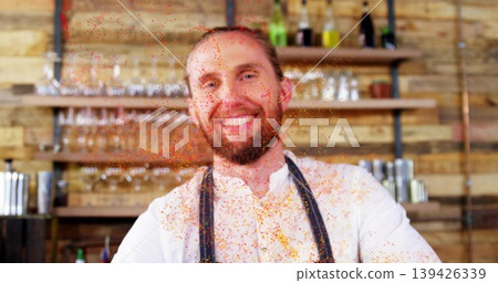 Smiling bartender in shirt and apron standing behind counter at bar, tossing orange spice particles Smiling bartender in shirt and apron standing behind counter at bar, tossing orange spice particles 139426339