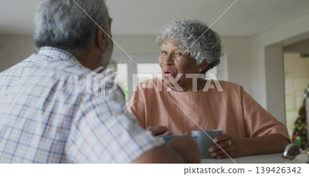 Chatting couple wearing salmon blouse at kitchen counter, holding coffee mugs next to eyeglasses 139426342