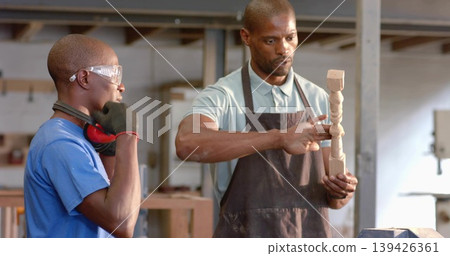 Showing mentor guiding apprentice examining wooden spindle at woodshop bench, with safety goggles Showing mentor guiding apprentice examining wooden spindle at woodshop bench, with safety goggles 139426361