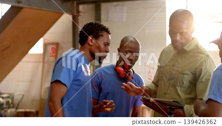 Examining tablet man in olive work shirt guiding blue tshirt workers in workshop with earmuffs Examining tablet man in olive work shirt guiding blue tshirt workers in workshop with earmuffs 139426362