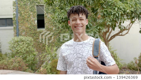 Smiling teen boy standing in campus courtyard carrying denim backpack and wearing patterned T-shirt Smiling teen boy standing in campus courtyard carrying denim backpack and wearing patterned T-shirt 139426369