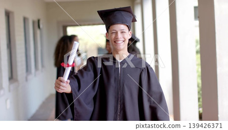 Walking students wearing black gowns and mortarboard caps down campus corridor, holding diplomas Walking students wearing black gowns and mortarboard caps down campus corridor, holding diplomas 139426371