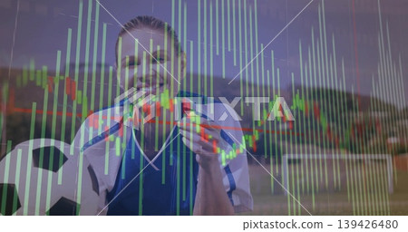 Smiling soccer player in sports jersey holding soccer ball biting medal on grass with chart overlay Smiling soccer player in sports jersey holding soccer ball biting medal on grass with chart overlay 139426480