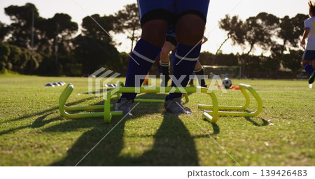 Soccer player feet stepping over neon agility hurdles in practice field, with cones and soccer ball 139426483