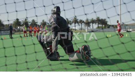 Kneeling field hockey goalkeeper wearing helmet and leg guards defending net on turf, with cones Kneeling field hockey goalkeeper wearing helmet and leg guards defending net on turf, with cones 139426485