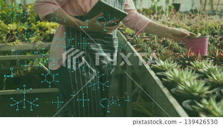 Technician wearing apron lifting pink pot and using tablet in greenhouse, with formula graphics 139426530