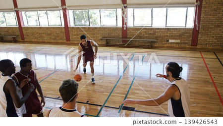 Dribbling adult male in maroon uniform moving across school gym court, holding orange basketball 139426543