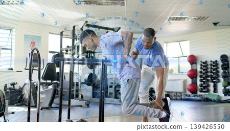 Doing dip client leaning forward over dip bars in gym, trainer supporting ankle wearing gym gear Doing dip client leaning forward over dip bars in gym, trainer supporting ankle wearing gym gear 139426550