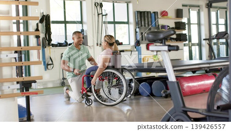 Kneeling therapist in green polo helping manual wheelchair client in rehab gym by bike, copy space Kneeling therapist in green polo helping manual wheelchair client in rehab gym by bike, copy space 139426557