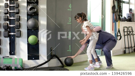 Supporting coach in sportswear guiding child reaching for med ball in gym, height marks, copy space 139426596