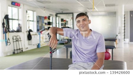 Sitting man in lavender shirt, gray shorts holding black forearm crutch on padded table, clinic 139426600