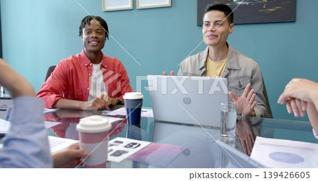 Discussing two colleagues in red button up and denim jacket at glass conference table, with laptop Discussing two colleagues in red button up and denim jacket at glass conference table, with laptop 139426605