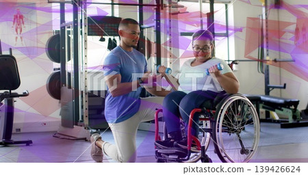 Spotting trainer guiding client in tee doing curls in manual wheelchair at gym, holding weights 139426624