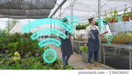 Inspecting two workers wearing aprons checking plants using tablet in nursery with cyan WiFi icons Inspecting two workers wearing aprons checking plants using tablet in nursery with cyan WiFi icons 139426642
