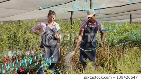 Watering and checking nursery staff in shade house with green hose and tablet, striped aprons Watering and checking nursery staff in shade house with green hose and tablet, striped aprons 139426647