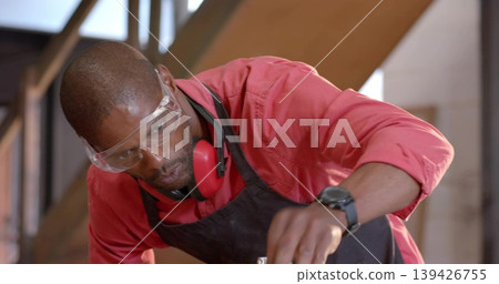 Leaning mature craftsman examining wooden plank in carpentry workshop, with goggles and earmuffs Leaning mature craftsman examining wooden plank in carpentry workshop, with goggles and earmuffs 139426755