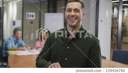 Smiling man wearing dark green shirt standing by wooden table in meeting room, reviewing papers 139426780