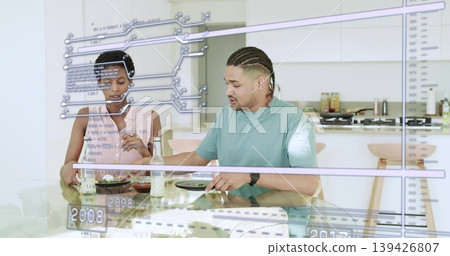 Couple eating at glass dining table with bottles in modern kitchen, showing digital overlays Couple eating at glass dining table with bottles in modern kitchen, showing digital overlays 139426807
